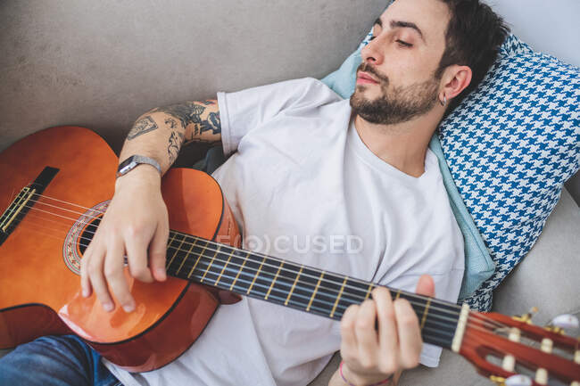Young bearded man lying on sofa, playing acoustic guitar — Stock Photo