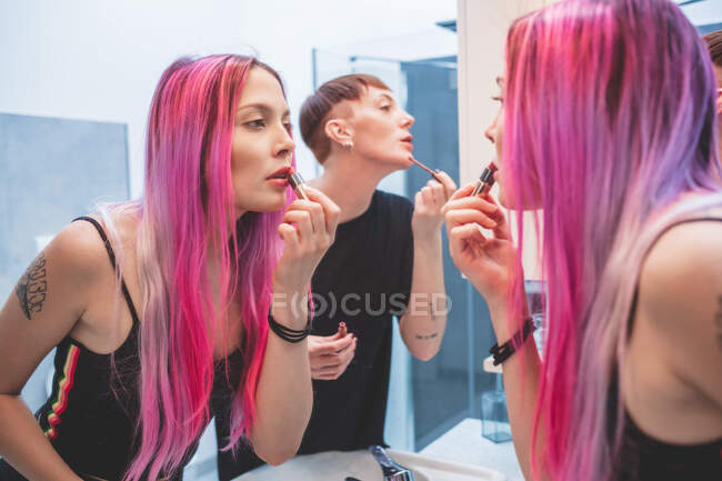 Mujer joven con el pelo largo y rosa y mujer con el pelo rojo corto de pie delante del espejo, aplicando lápiz labial en los labios - foto de stock