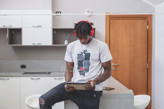 Молодий чоловік з короткими замками, сидячи на кухні, носить червоні навушники і тримає цифровий планшет, дивлячись на екран — стокове фото