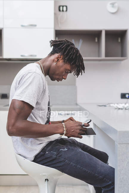Young man with short dreadlocks sitting on bar stool in kitchen, using mobile phone, smartphone in hands — Stock Photo