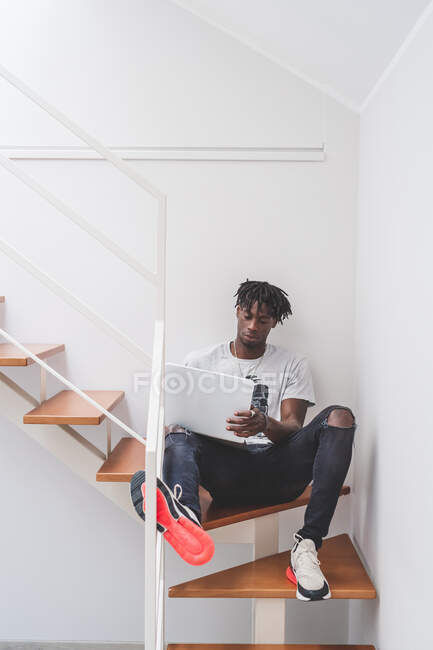 Junger Mann mit kurzen Dreadlocks sitzt im Treppenhaus und tippt auf Laptop-Notizbuch — Stockfoto