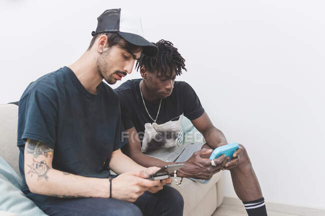 Deux jeunes hommes assis sur le canapé, vérifiant leurs téléphones portables, smartphones dans les mains — Photo de stock