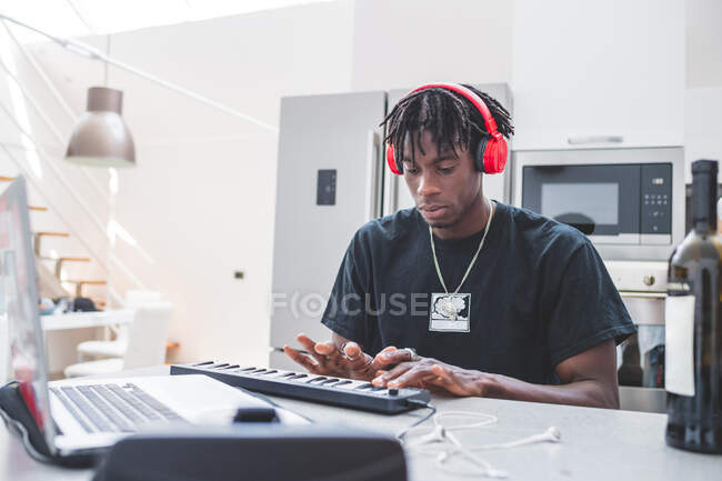 Joven hombre afroamericano con rastas cortas sentado en una mesa, con auriculares, tecleando en el teclado - foto de stock
