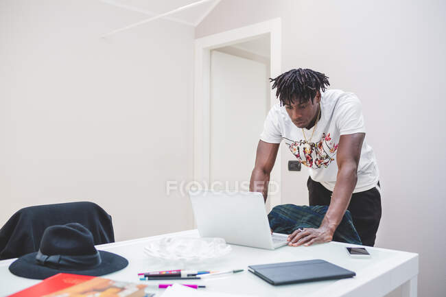 Young African american man with short dreadlocks standing at table, looking at laptop notebook — Stock Photo