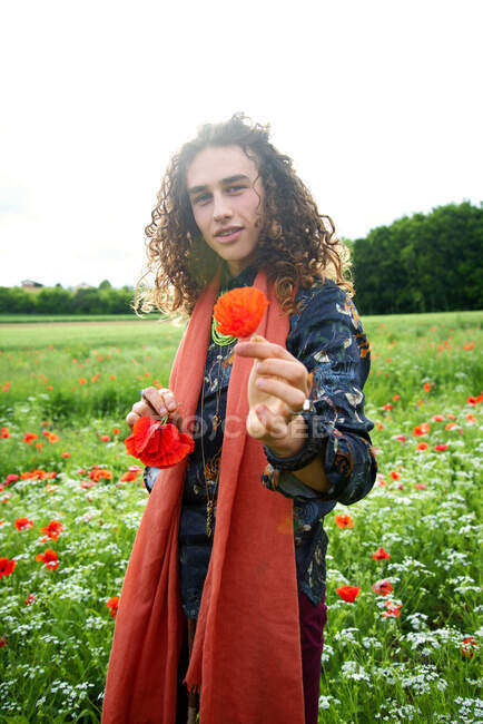 Portrait of young man with long brown curly hair in poppies meadow, showing flower on camera — Stock Photo