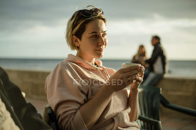 Portrait of smiling young blond woman leaning against a car parked by the sea at dusk — Stock Photo