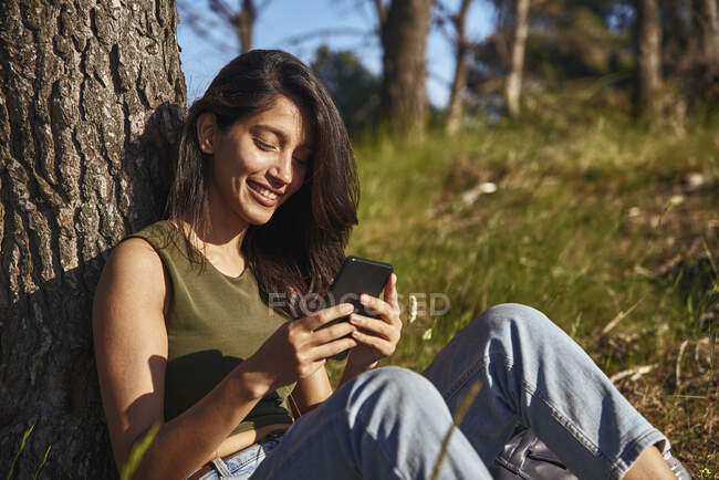 Портрет молодой женщины, сидящей под деревом в лесу, улыбающейся и просматривающей мобильный телефон — стоковое фото
