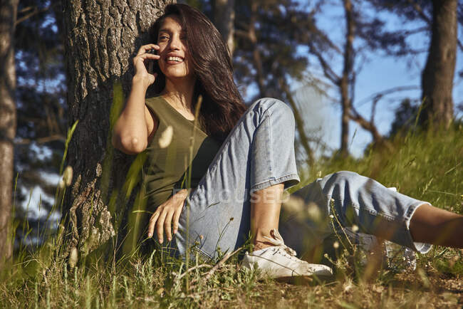 Portrait de jeune femme aux longs cheveux bruns assise sous un arbre dans une forêt, parlant sur téléphone portable — Photo de stock