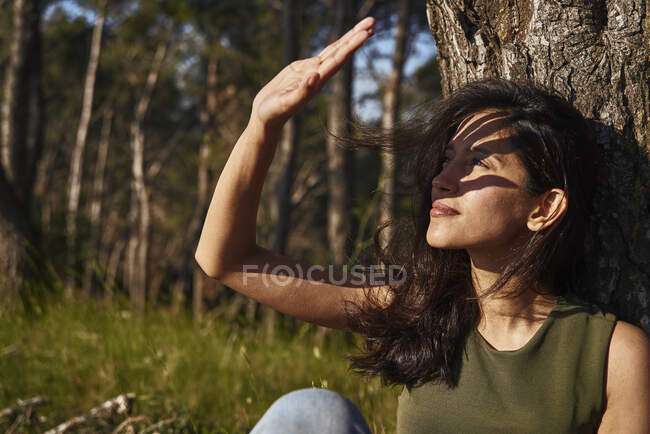 Portrait of young woman sitting under a tree in a forest, shading her eyes from the sun — Stock Photo
