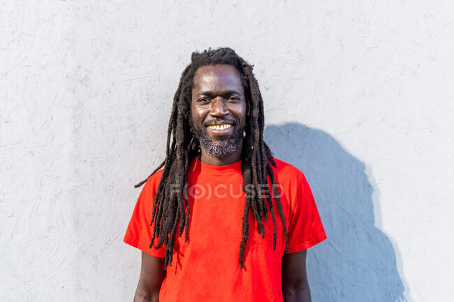 Portrait of Black man with dreadlocks wearing red T-Shirt, standing in front of white wall, smiling at camera. — Stock Photo