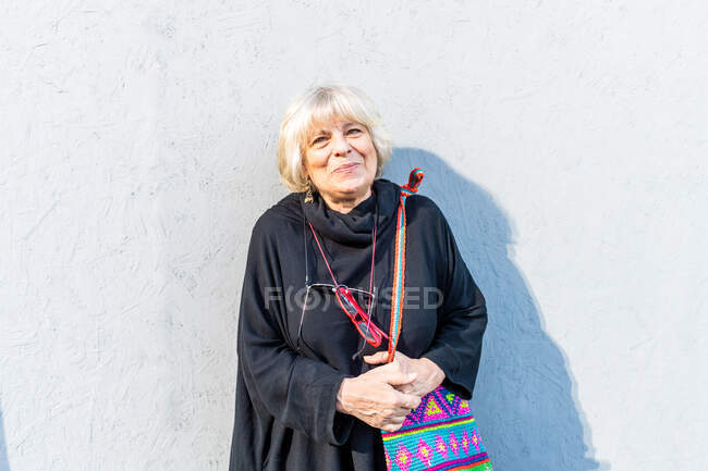 Портрет пожилой женщины с светлыми волосами в черном топе, стоящей перед белой стеной, улыбающейся в камеру. — стоковое фото