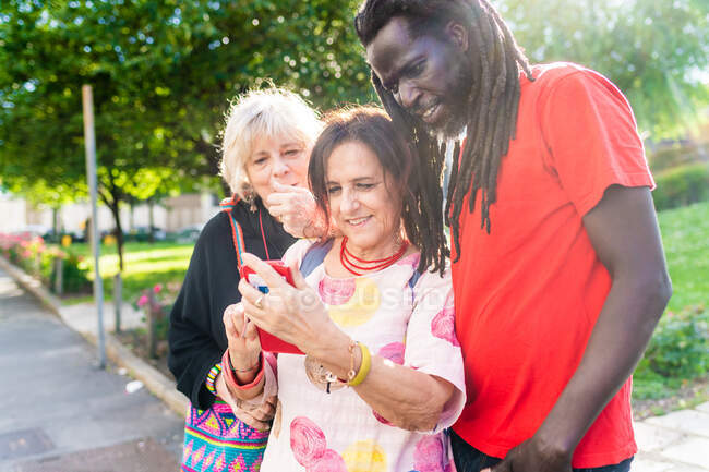 Hombre negro con rastas y dos mujeres caucásicas de pie en el puente, mirando el teléfono móvil. - foto de stock