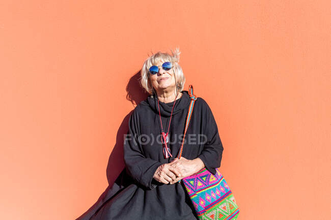 Ritratto di donna anziana con capelli biondi e occhiali da sole davanti alla parete arancione. — Foto stock