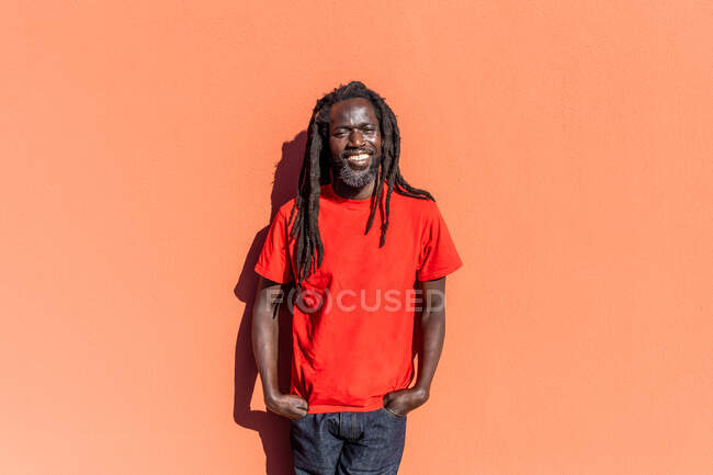 Retrato del hombre negro con rastas de pie frente a la pared naranja, sonriendo a la cámara. - foto de stock