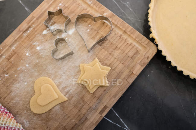High angle close up of cookie cutters and cut out dough in star and heart shapes on wooden cutting board. — Stock Photo