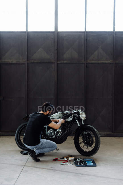 Jovem ajoelhado para fazer manutenção em motocicleta vintage em armazém vazio — Fotografia de Stock