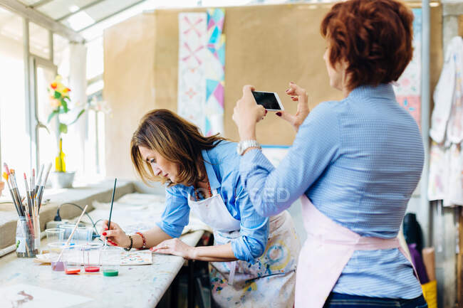 Pintura de artista en tela en un estudio creativo, mujer madura fotografiándola con un teléfono inteligente - foto de stock