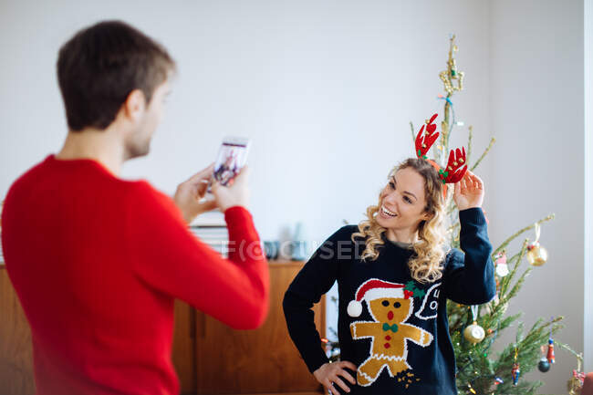 Homem tirando foto de mulher na frente da árvore de Natal em casa — Fotografia de Stock