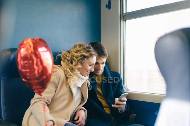 Couple avec ballon en forme de coeur en utilisant un smartphone à l'intérieur du train — Photo de stock