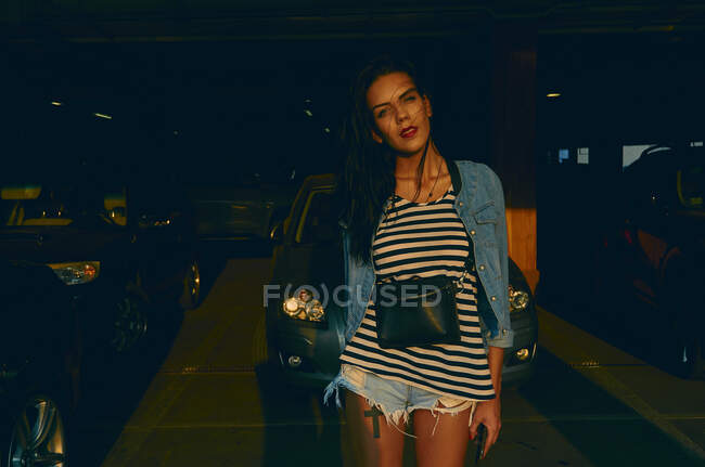 Портрет женщины с длинными каштановыми волосами, в горячих штанах, стоящей ночью на парковке. — стоковое фото