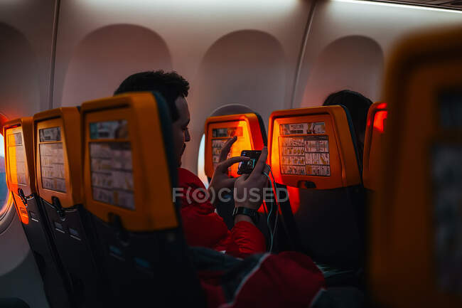 Over the shoulder view of man sitting in passenger plane, using mobile phone. — Stock Photo