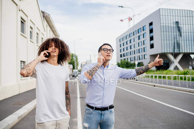 Two men with tattooed arms standing on sidewalk, using mobile phones, hitchhiking. — Stock Photo