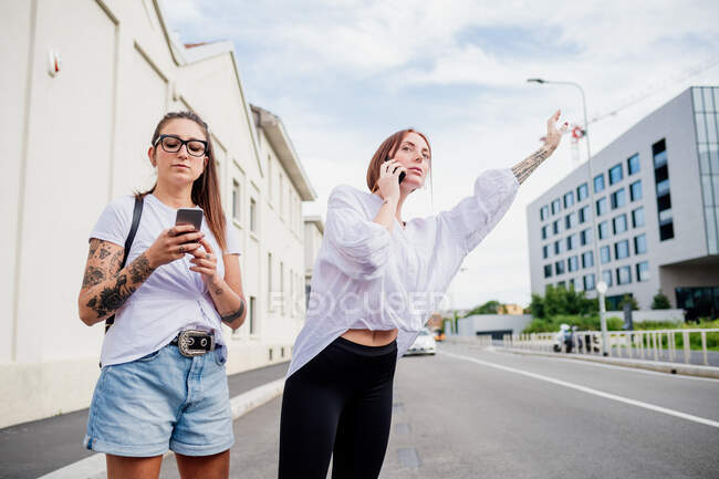Two women with tattooed arms standing on sidewalk, using mobile phones, hailing taxi. — Stock Photo