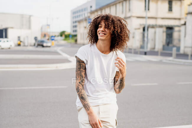 Homem sorridente com braços tatuados e cabelos longos e castanhos encaracolados em pé em uma rua. — Fotografia de Stock