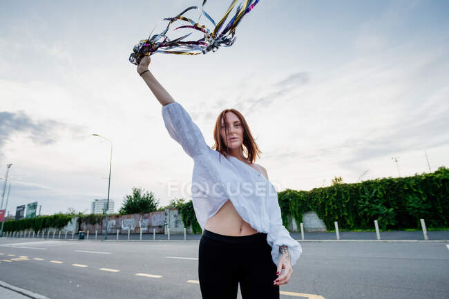 Portrait of woman with long red hair, standing on street, holding streamers in raised hand. — Stock Photo
