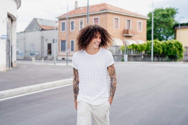 Smiling man with tattooed arms and long brown curly hair walking down street. — Stock Photo