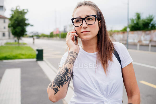 Portrait de femme aux longs cheveux bruns et au bras tatoué, portant un T-shirt blanc et des lunettes, utilisant un téléphone portable. — Photo de stock