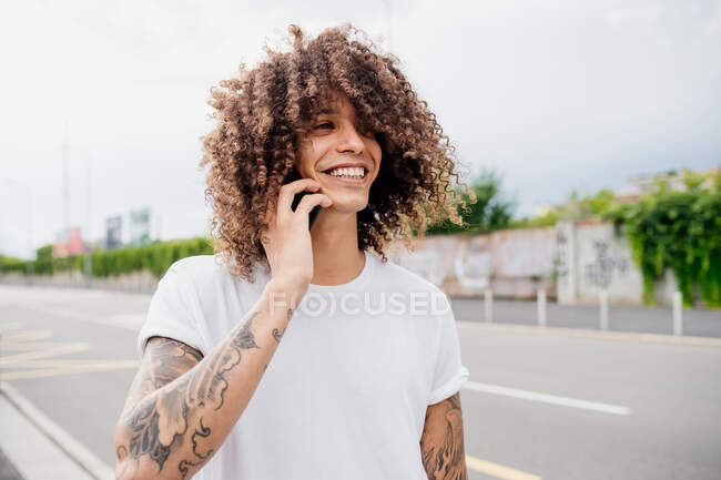 Retrato de homem com braços tatuados e cabelos longos e castanhos encaracolados, usando telefone celular. — Fotografia de Stock