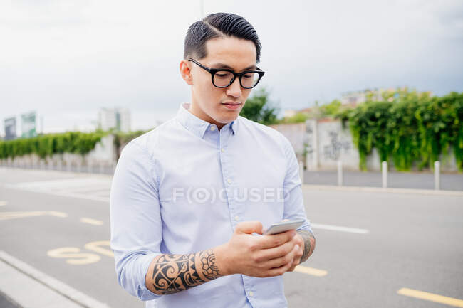 Retrato de hombre con brazo tatuado, con camisa y gafas de color azul claro, usando teléfono móvil. - foto de stock