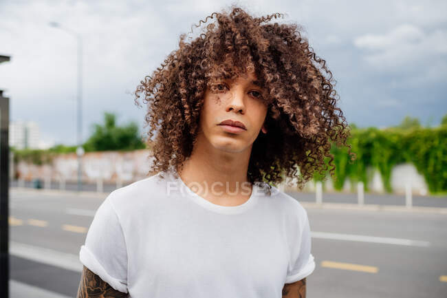 Retrato del hombre con el pelo largo y rizado marrón, vistiendo una camiseta blanca, mirando a la cámara. - foto de stock