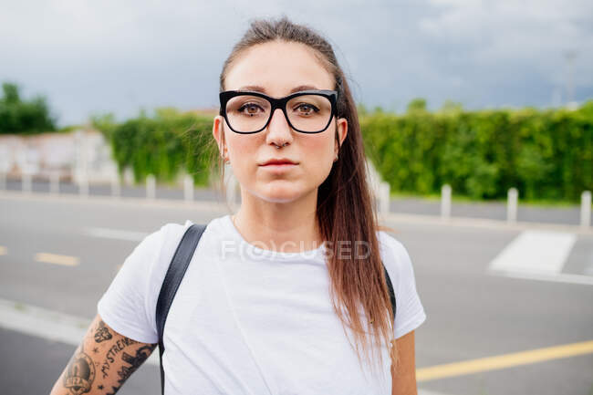 Retrato de mulher com cabelos castanhos longos e braço tatuado, vestindo camiseta branca e óculos, olhando para a câmera. — Fotografia de Stock