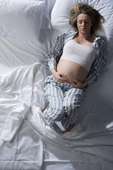 Portrait of heavily pregnant woman lying on bed, cradling stomach. — Stock Photo