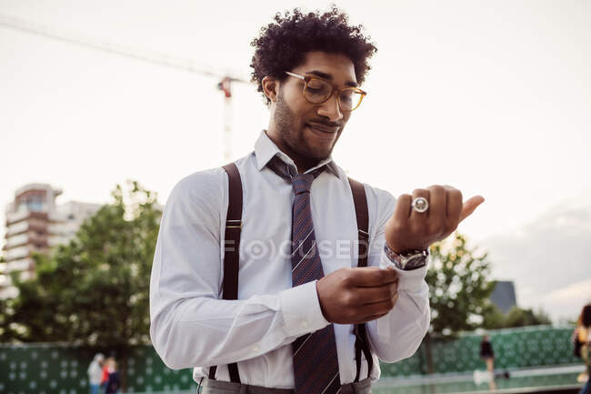 Retrato de hombre de negocios con gafas, camisa blanca, corbata marrón y tirantes, ajustando sus puños. - foto de stock