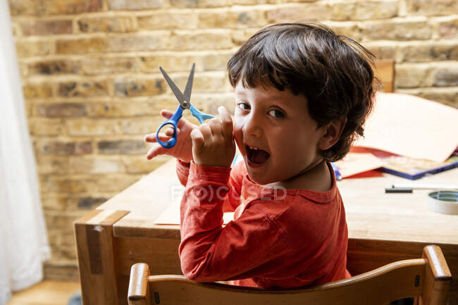 Niño con el pelo castaño sentado en la mesa, sosteniendo un par de tijeras, sonriendo a la cámara. - foto de stock