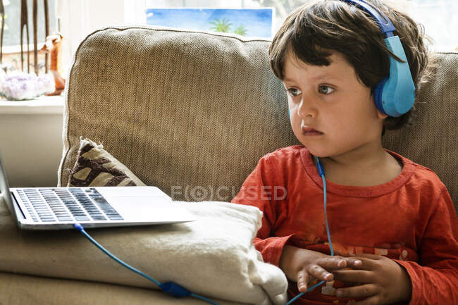 Молодий хлопчик з коричневим волоссям носить сині навушники, спостерігаючи за екраном ноутбука . — стокове фото