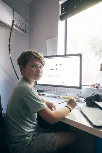 Niño sentado en su escritorio delante de la computadora, mirando a la cámara. - foto de stock