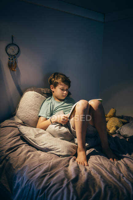 Niño sentado en la cama en su habitación, leyendo. - foto de stock