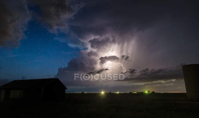 Многочисленные удары молнии из облака в облака видны вдоль сухого участка ночью, со звездами на заднем плане над фермерскими зданиями — стоковое фото