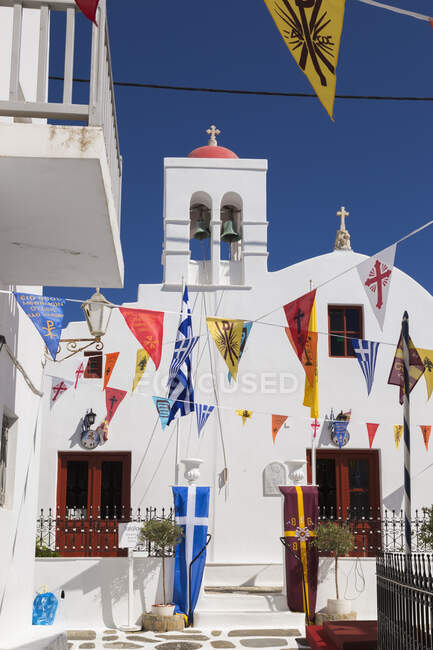 Chiesa greco-ortodossa e bandiere colorate appese sopra stretto vicolo nella città di Mykonos. — Foto stock