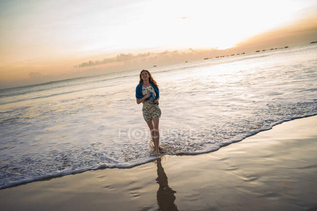 Mujer adulta que camina en el mar al atardecer, Bahía Jimbaran, Bali, Indonesia - foto de stock