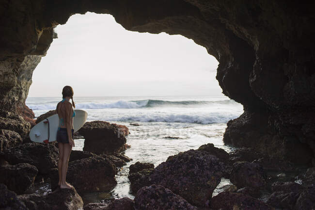 Donna in piedi su rocce vicino alla baia, tenendo tavola da surf, Nusa Ceningan, Indonesia — Foto stock