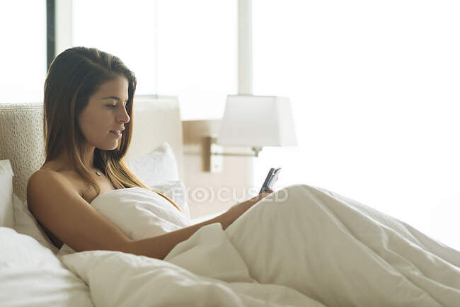 Young woman sitting up in hotel bed reading smartphone texts — Stock Photo
