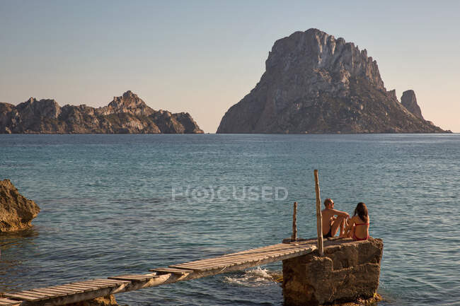Tourist couple sitting on pier looking over to Es Vedra, Ibiza, — Stock Photo