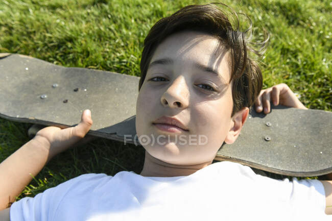 Menino com cabelo castanho deitado na grama, cabeça descansando no skate, olhando para a câmera — Fotografia de Stock