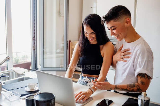 Jovem casal lésbico de pé na cozinha, usando laptop. — Fotografia de Stock