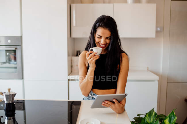 Молода жінка з довгим коричневим волоссям стоїть на кухні, дивлячись на цифровий планшет — стокове фото